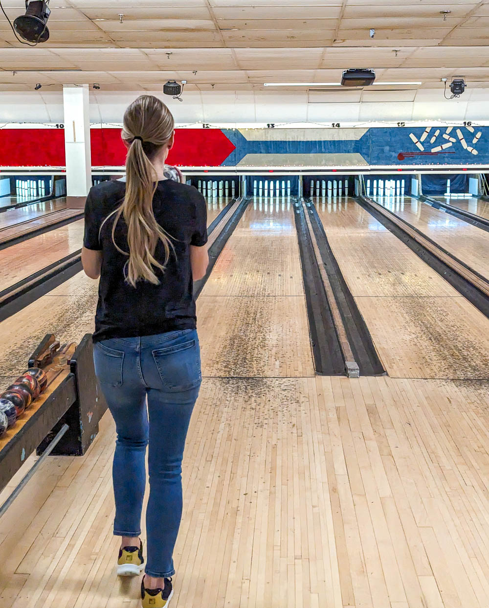 woman in jeans and black tshirt getting ready to roll a ball down a long wooden lane